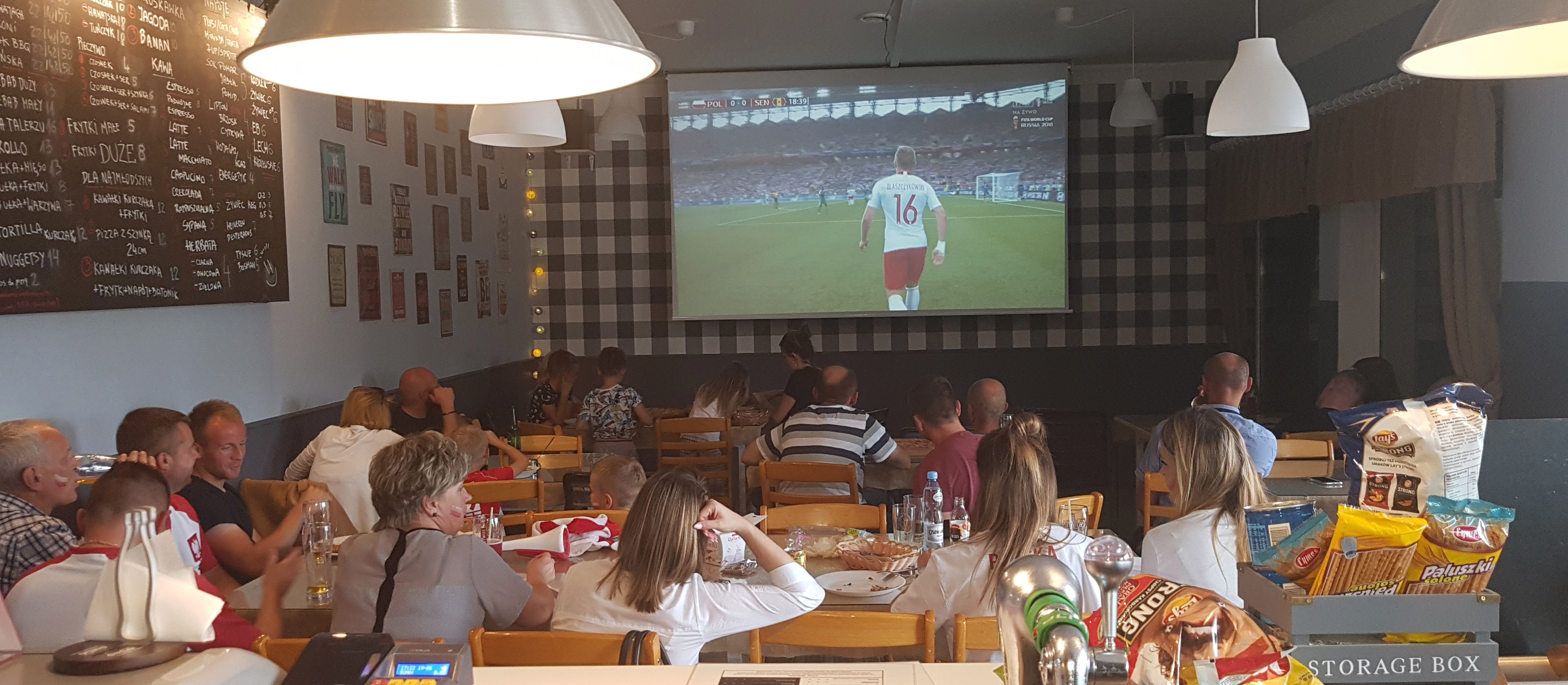 Zdjęcie przedstawia wnętrze pizzerii, w której siedzi ponad 20 osób, w tym małe dzieci. 
            Wszyscy oglądają mecz reprezentacji Polski na ponad stucalowym ekranie.